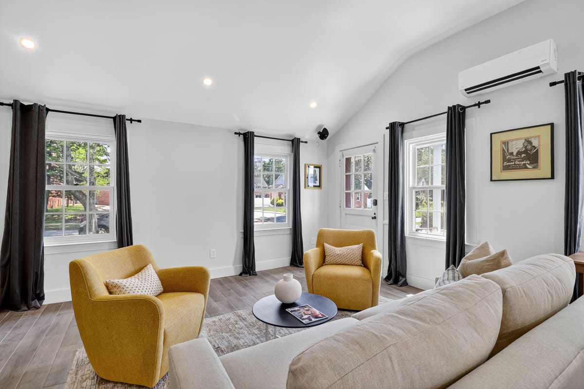 Living room from the far corner, with mustard swivel chairs, a cream sofa, a round black coffee table and windows dressed in black linen.