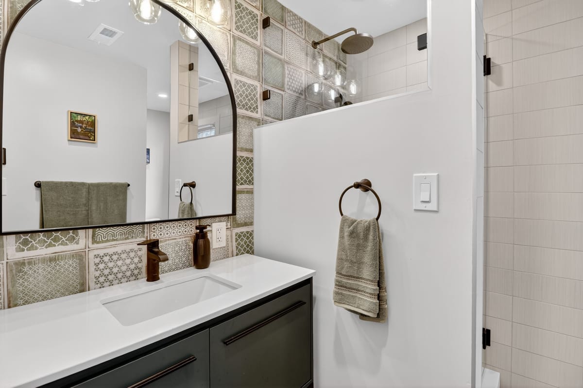 Bathroom with a black vanity, white quartz top, an arched black mirror and green patterned tile in the shower.