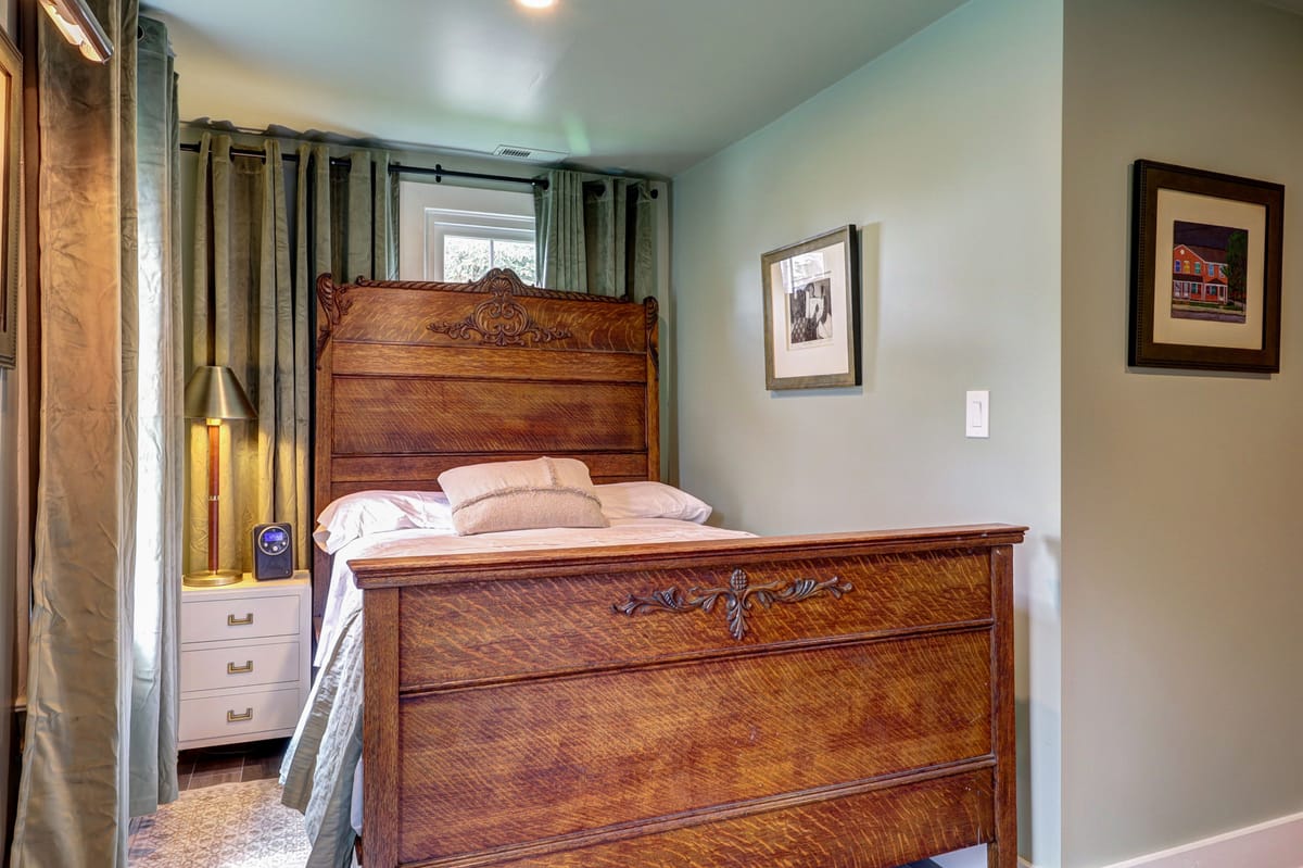 Bedroom with a carved antique wooden bed against a sage wall, framed prints, a white nightstand and a pine barn door.