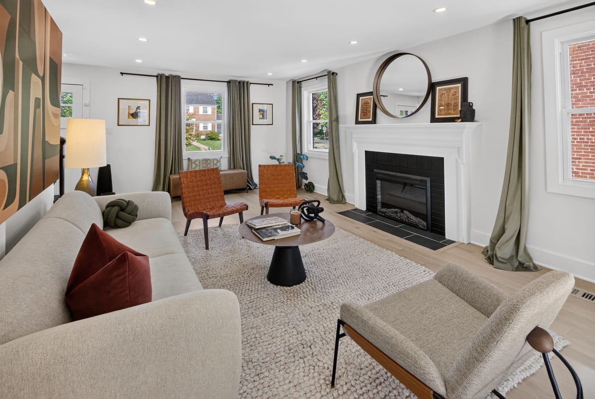 The finished living room, with a pale sectional, a low black side table, olive drapery and framed art stacked beside a round mirror above the fireplace.