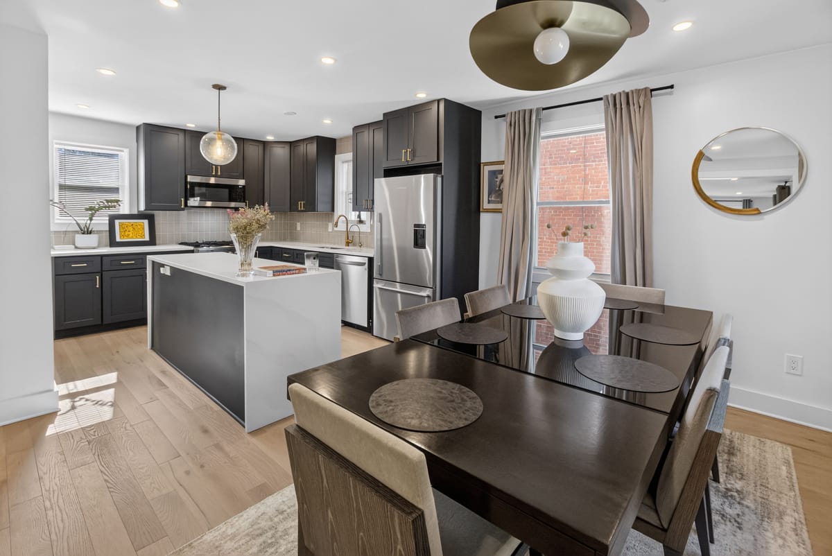 The finished kitchen and dining room, with black cabinetry, brass pulls, a stone island, a dark timber dining table and a sculptural brass pendant.