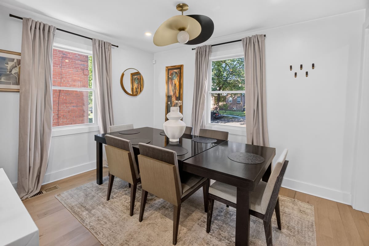 Dining room with a dark timber table, upholstered chairs, a round gilt mirror between two windows and floor-length linen curtains.