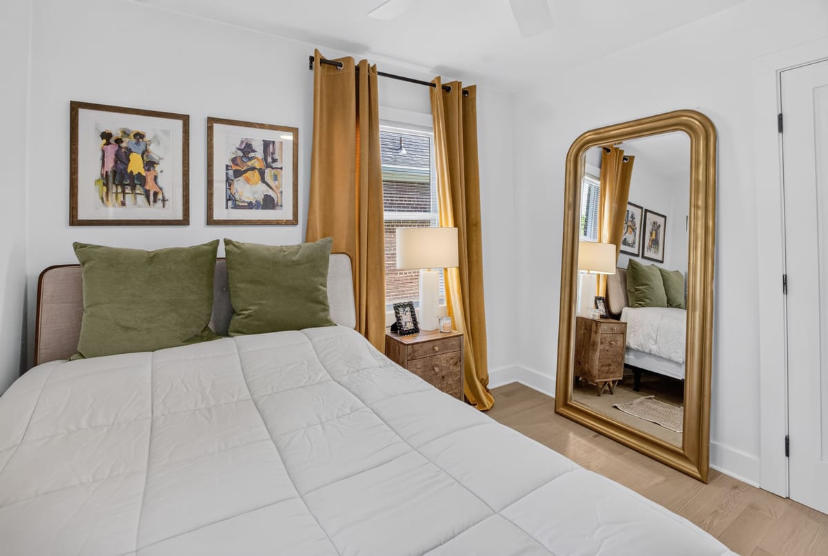 Bedroom with a white-dressed bed, olive velvet curtains, a tall gilt-framed mirror and a row of small framed prints.