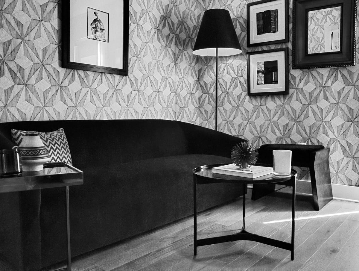 Black and white detail of a curved sofa against patterned wallpaper, with a gallery wall of framed prints and a slim floor lamp.