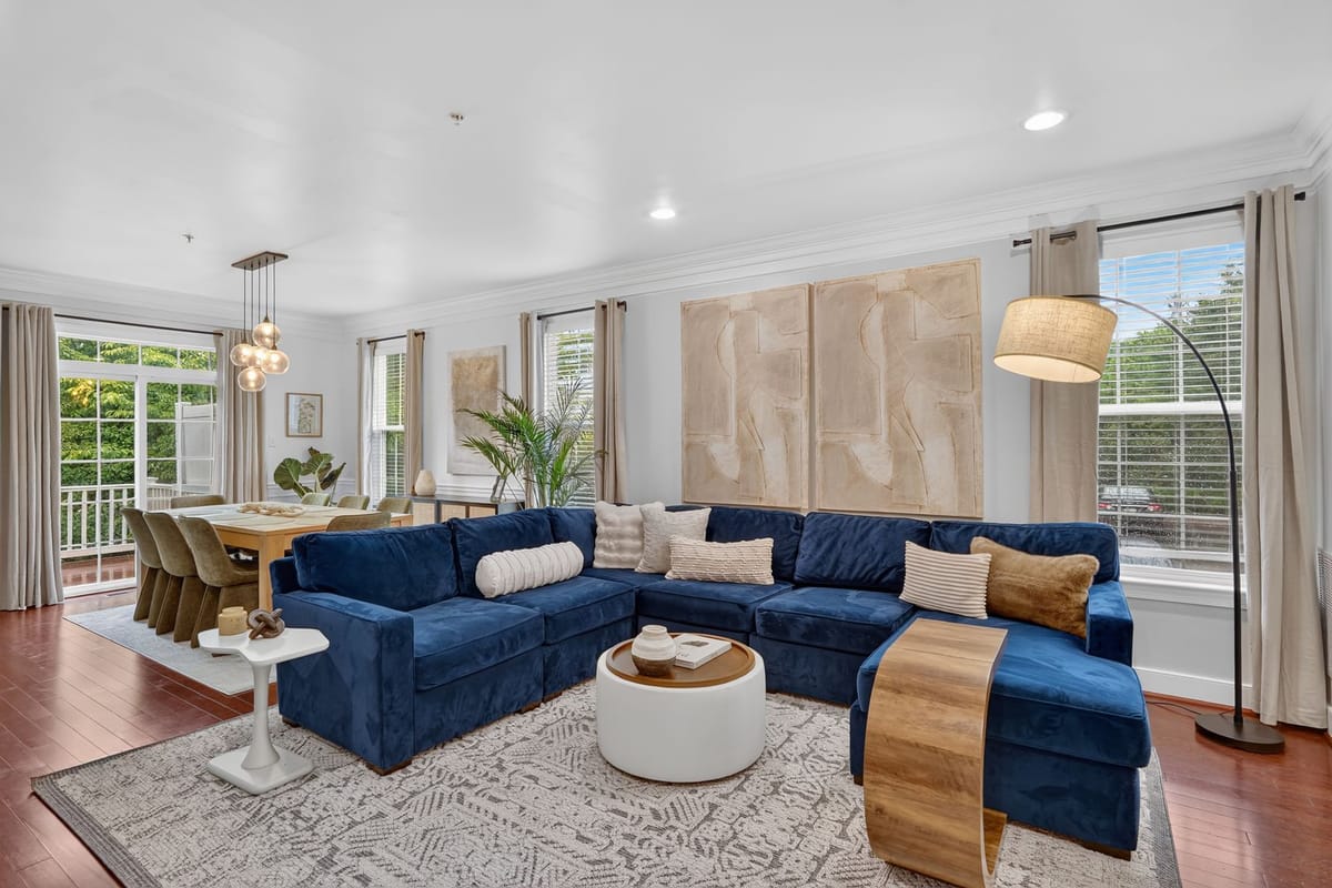 The finished living room with a deep navy velvet sectional, a round white ottoman, a pale patterned rug and tall plants at the windows.