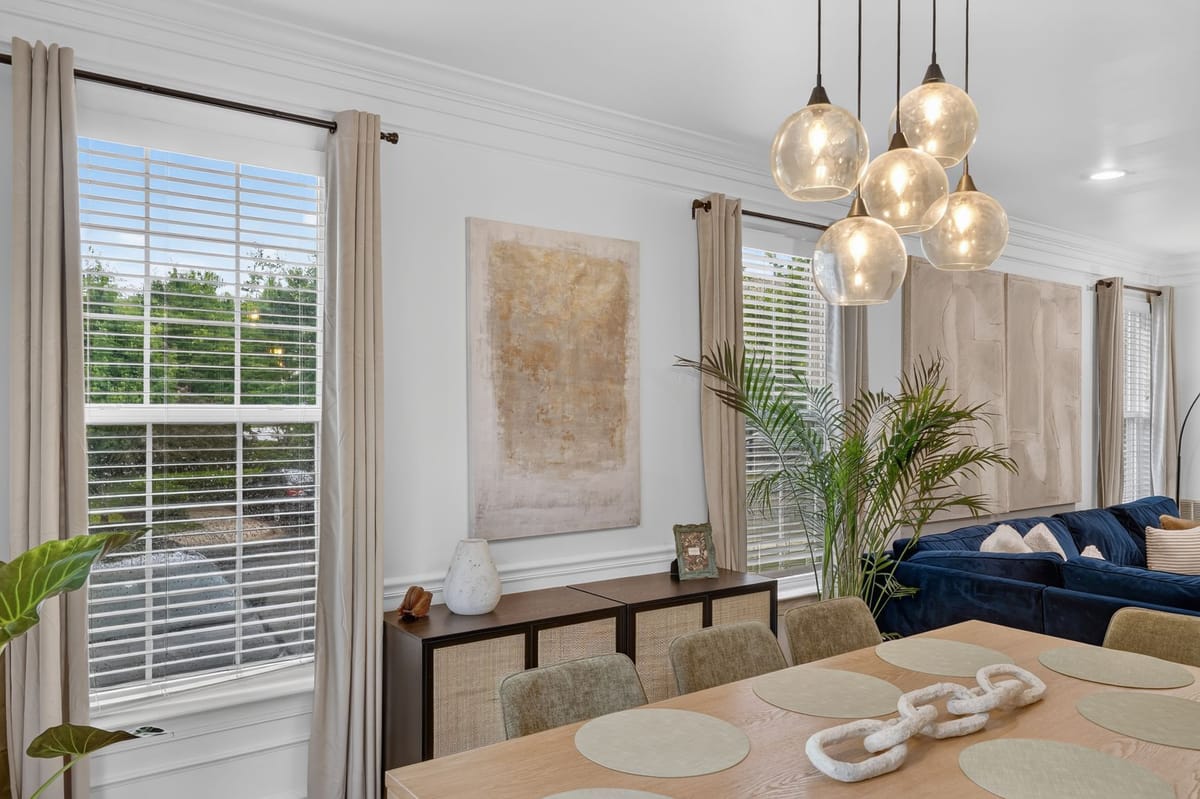 Living and dining rooms seen together, with the navy sectional at one end and the oak dining table at the other beneath layered linen curtains.