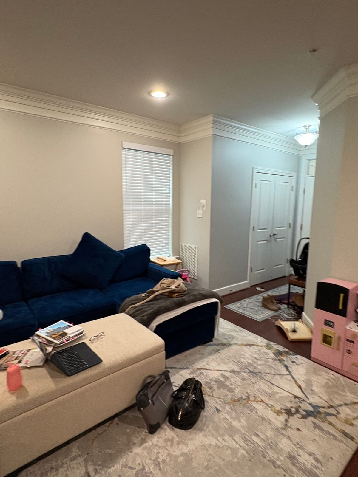 The living room as found, with a navy sofa, a cream storage ottoman and toys and papers across the rug.