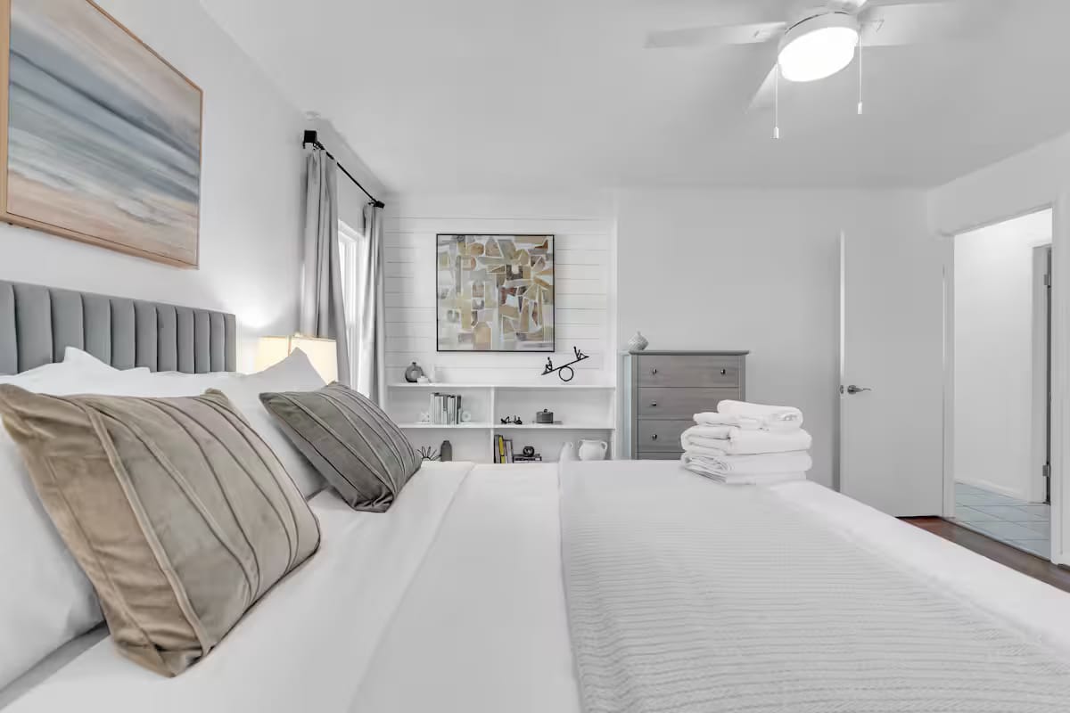 Guest bedroom with a channel-tufted grey headboard, layered white bedding, a folded throw and a small framed collage above the dresser.