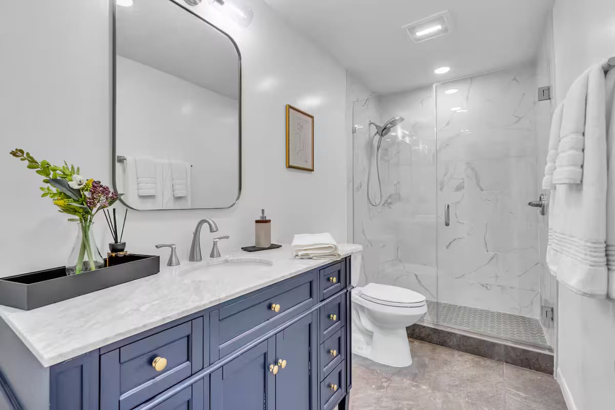 The finished bathroom with a navy vanity, brass fittings, a marble top, an arched black-framed mirror and a walk-in marble shower.