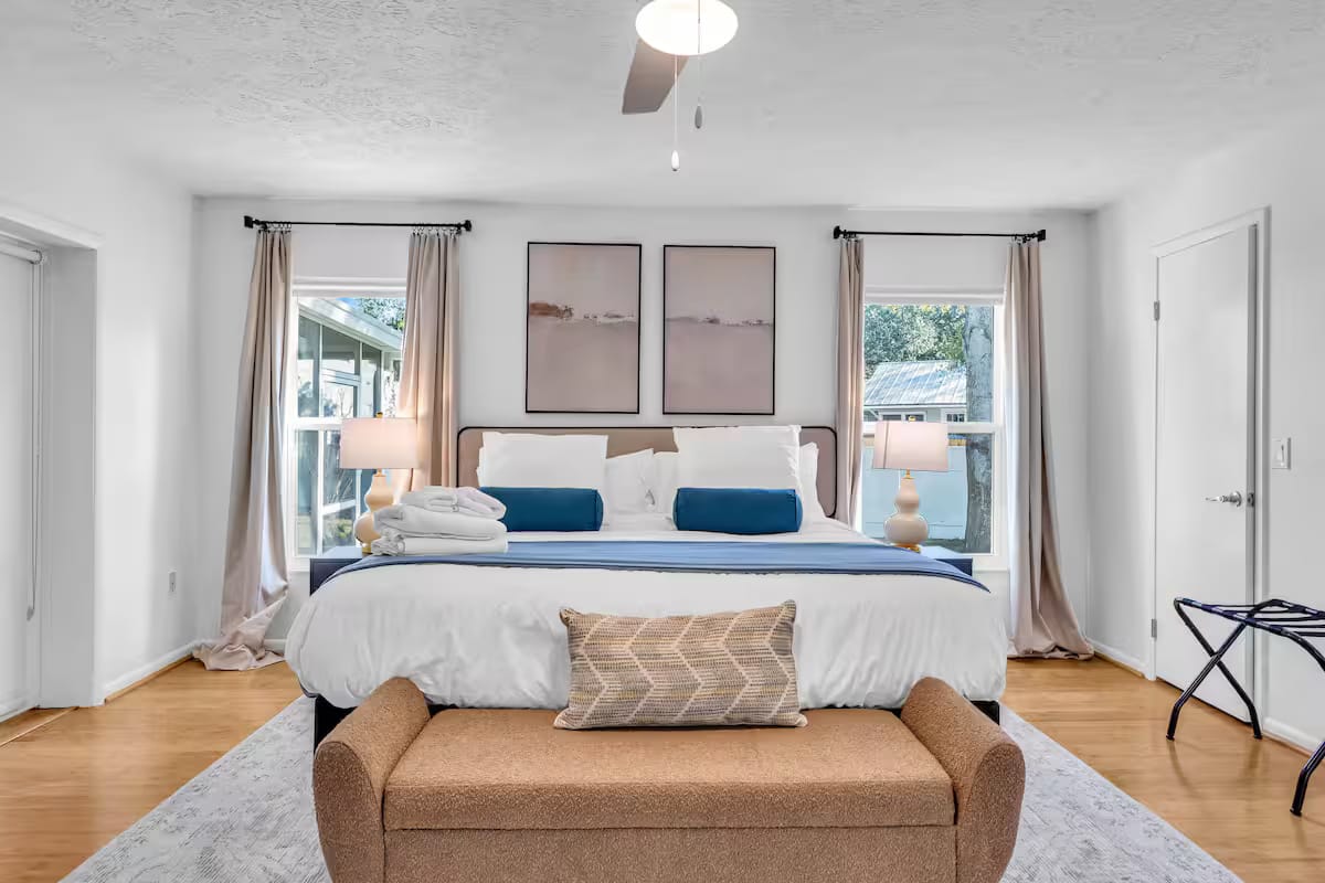 The finished bedroom with a denim-blue upholstered bed, white linens, a tan bench at the foot and a pair of soft landscape prints above the headboard.