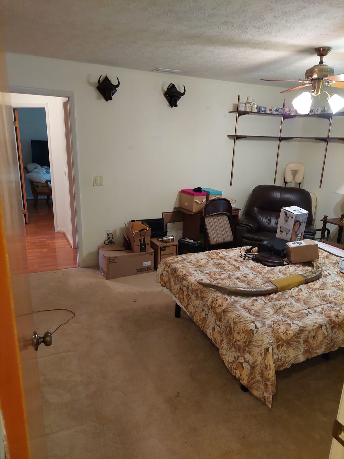 The bedroom as found, with a floral bedspread, a dark headboard and boxes stacked against the far wall under a ceiling fan.