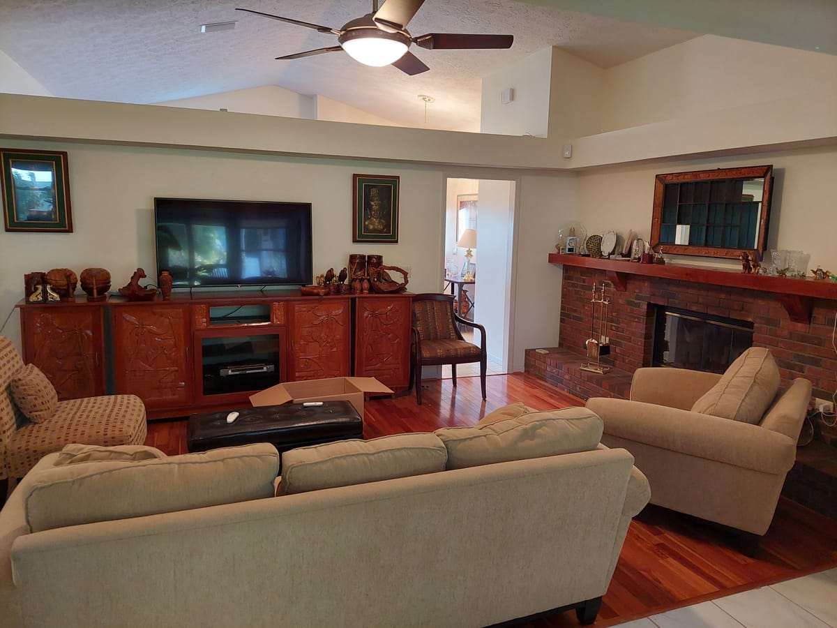 A family room as found, with a brick fireplace, a large timber media cabinet and patterned sofas.