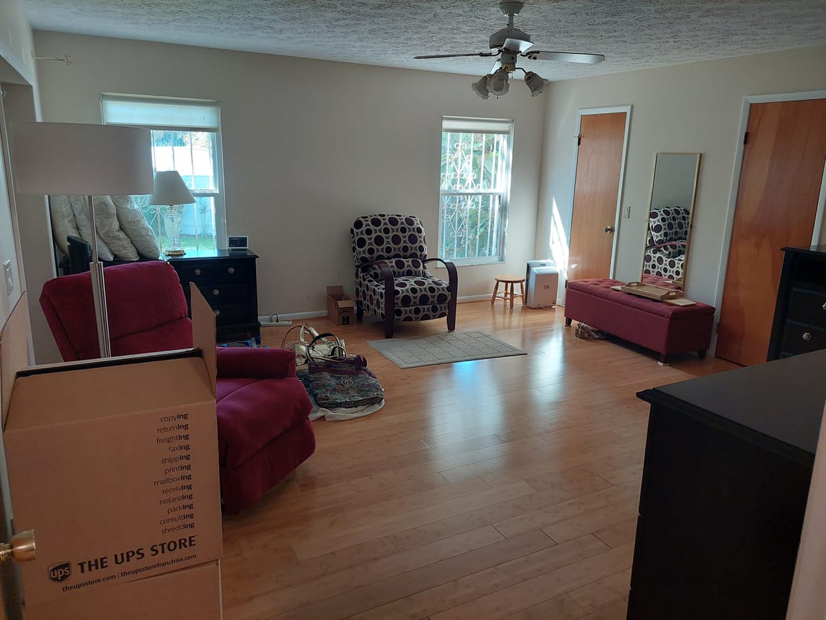 A living room as found, with moving boxes stacked beside a burgundy sofa and dark occasional chairs.